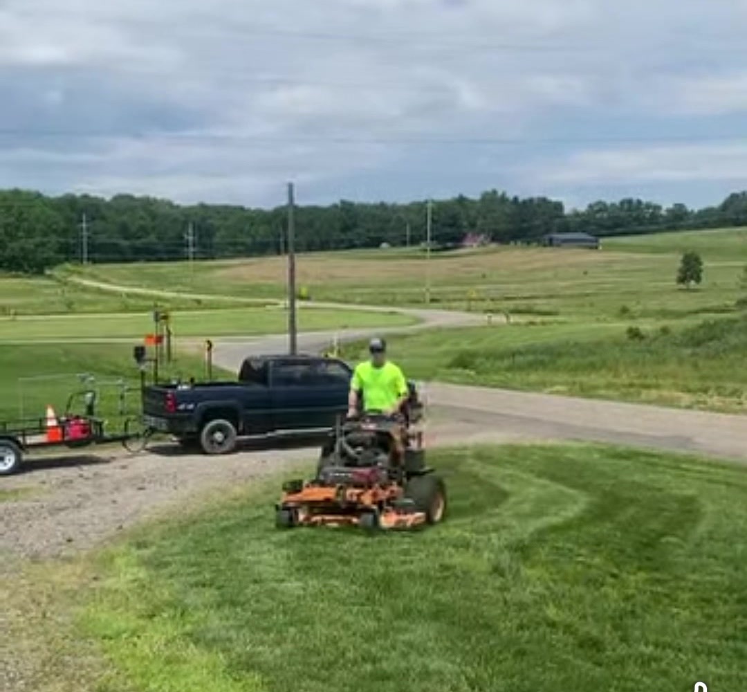A crew member in a neon shirt mowing with the truck behind